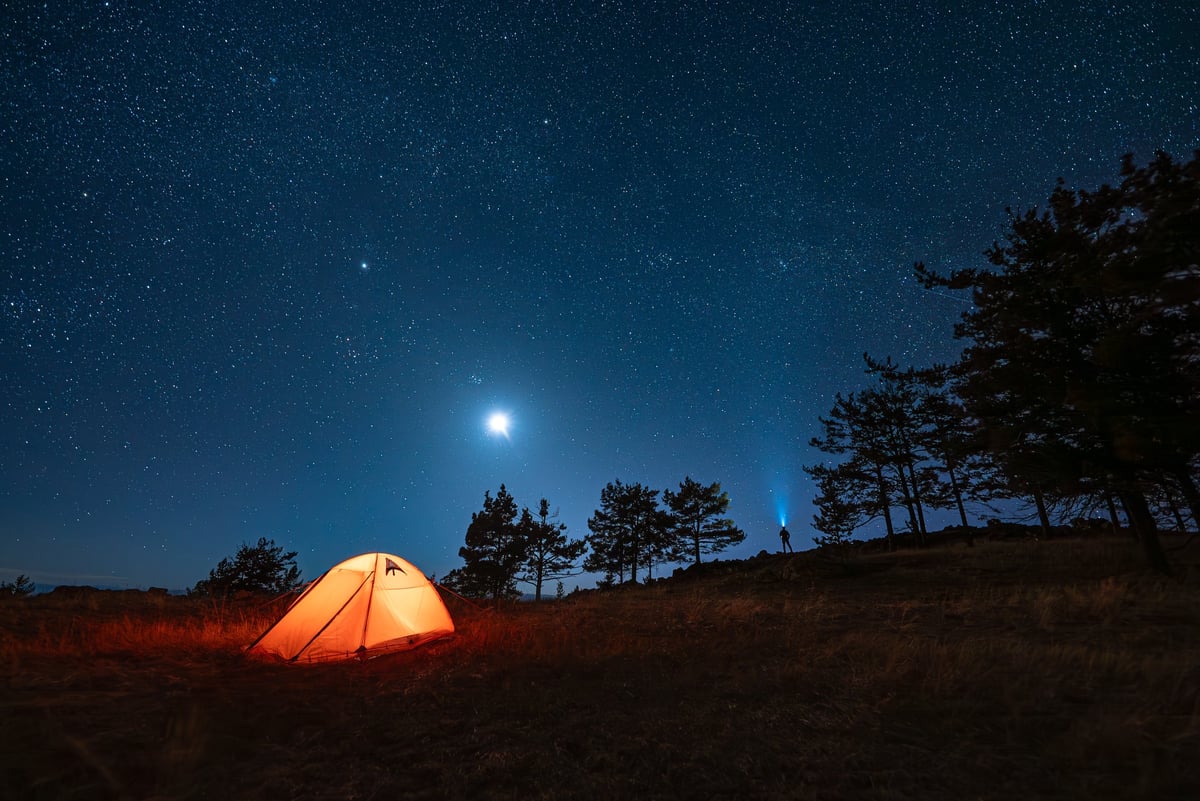 Small silhouette of alone hiker at his illuminated camp tent at starry night