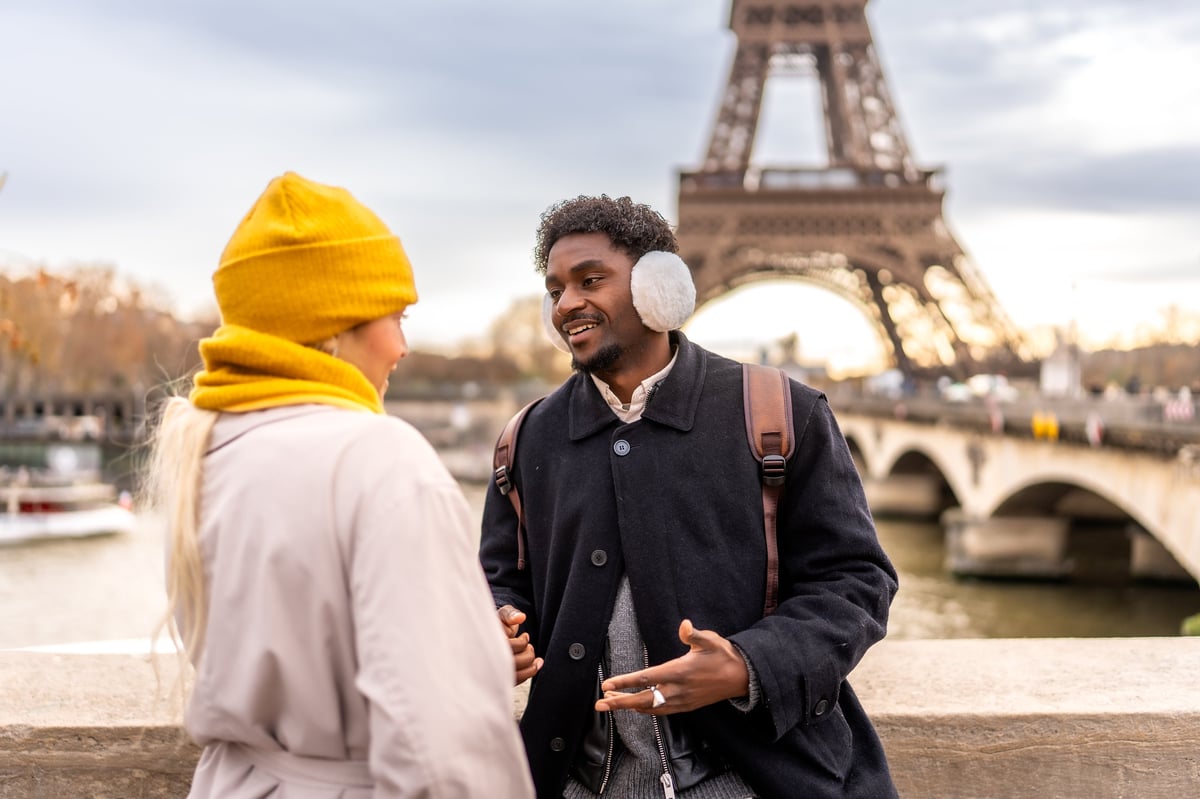 Young multiracial couple sharing conversation and smiles on a paris bridge, eiffel tower behind them, bundled in winter coats and scarves during a romantic city break
