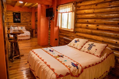Rustic log cabin bedroom with a double bed, wooden walls, warm lighting, and an adjacent bathroom view through the doorway