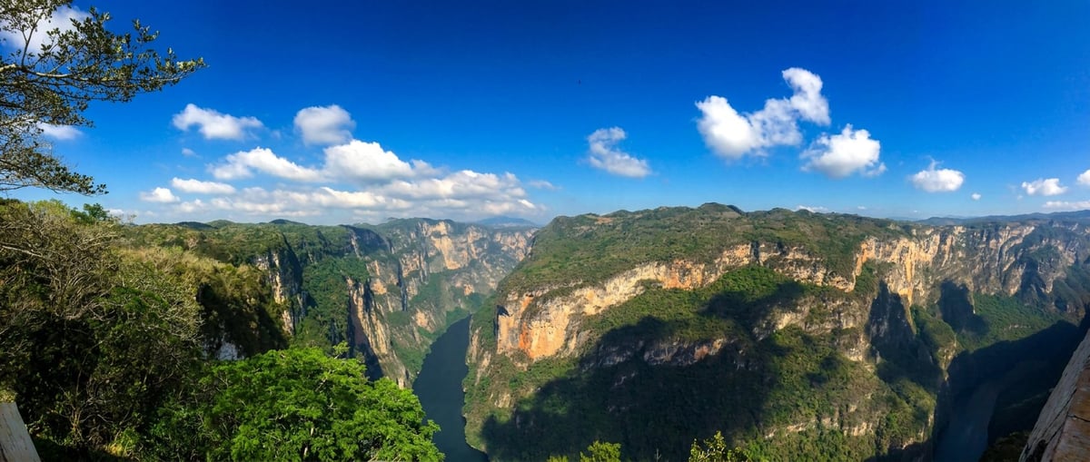 Panoramic view of a dramatic mountain canyon with steep rock cliffs, lush green valleys, and blue sky with white clouds