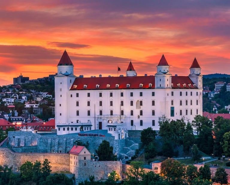 Bratislava Castle illuminated at sunset with dramatic orange and purple sky, surrounded by fortification walls and trees