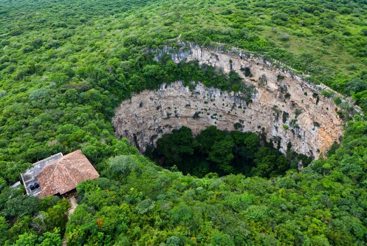 Aerial view of a large sinkhole surrounded by lush green forest with a small structure visible on the left