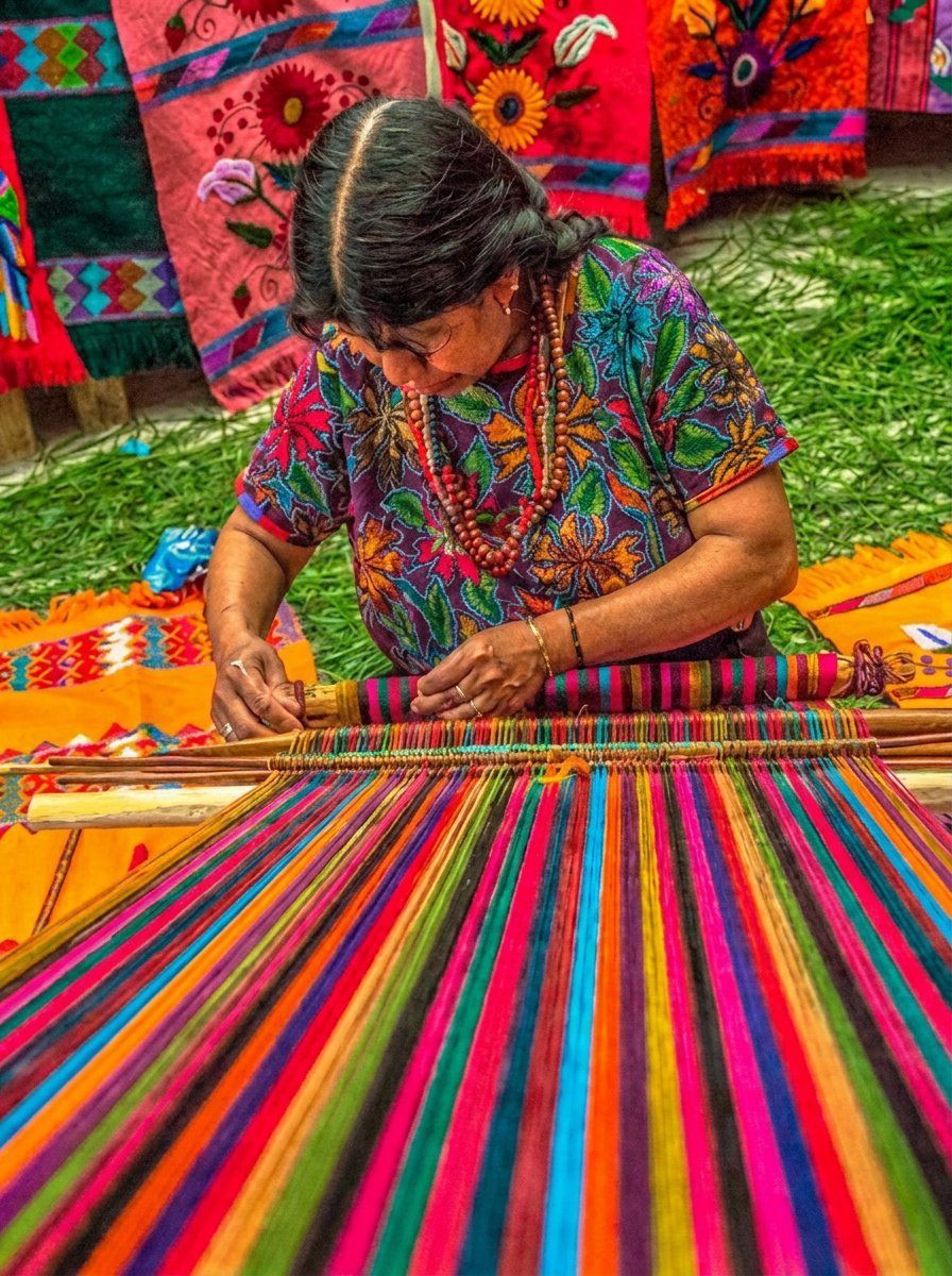 Woman weaving traditional colorful textiles on a loom with vibrant indigenous fabrics and designs displayed in background