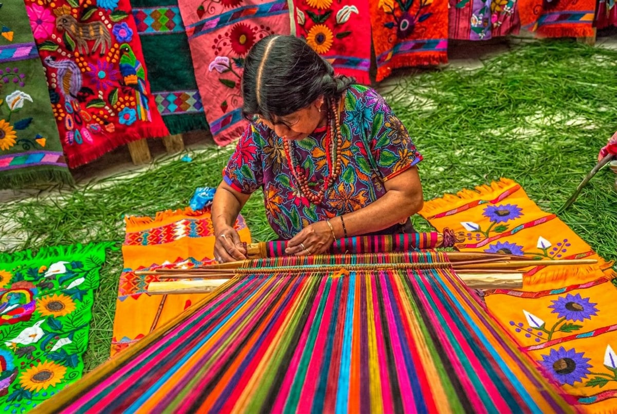 Artisan woman weaving colorful traditional textile on wooden loom outdoors with vibrant fabric displays in background