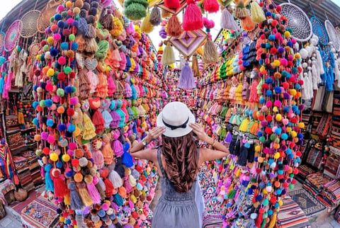 Person wearing white hat standing in colorful market stall surrounded by hanging pom-poms and yarn balls