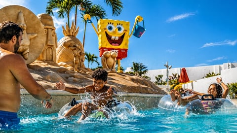 Niños salpicando en piscina con estatua de Bob Esponja y palmeras de fondo en parque acuático