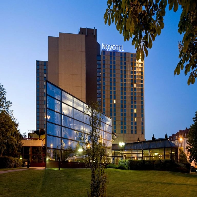 Novotel hotel exterior at dusk with illuminated modern glass entrance, brick tower, and landscaped courtyard under clear blue sky