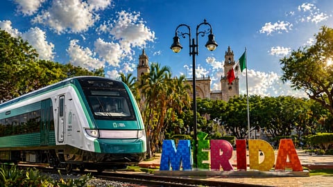 Tren moderno verde y blanco junto a señal colorida MÉRIDA con palmeras, farol y catedral al fondo