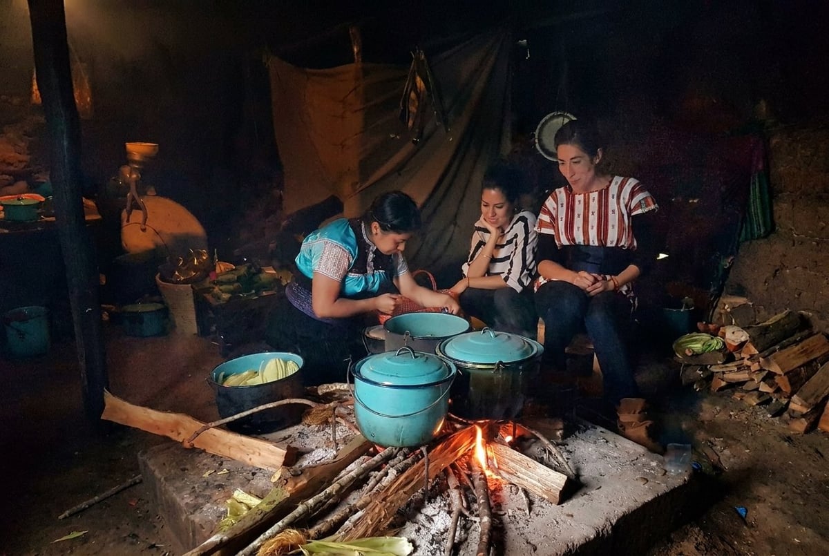 Family gathering around a campfire at night, cooking in blue pots over flames, with people seated on logs in casual clothing