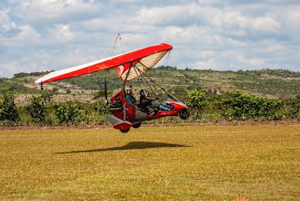 Red and white hang glider flying low over a rural field with hills and vegetation in the background