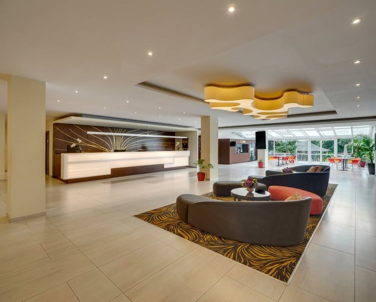 Modern hotel lobby with curved seating area, reception desk, bronze and gold ceiling elements, and floor-to-ceiling windows overlooking a garden