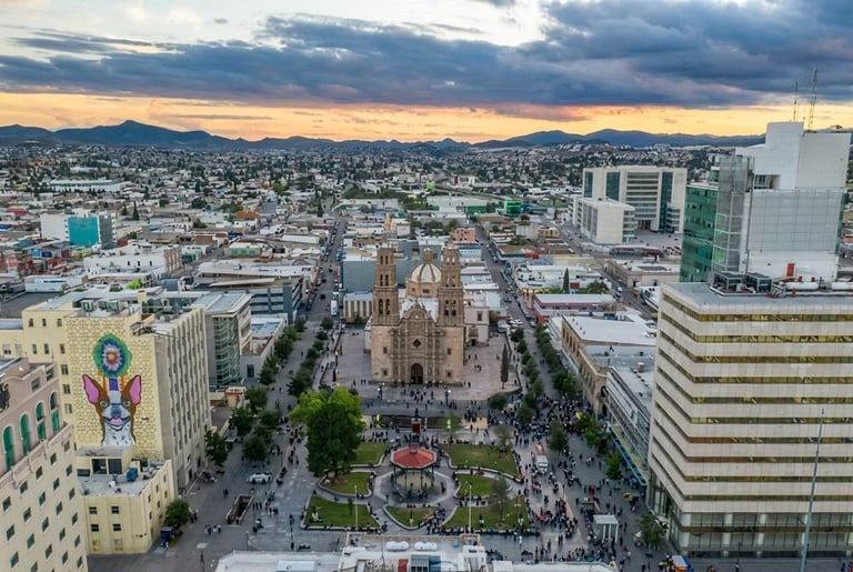 Aerial view of a cathedral in a city plaza surrounded by buildings at sunset with mountains in the background