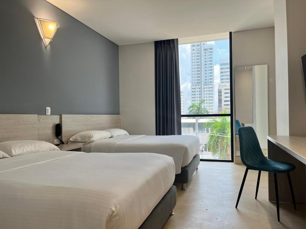 Modern hotel room with two twin beds, gray accent wall, floor-to-ceiling windows overlooking city skyline, and contemporary furnishings.
