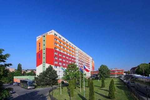 Modern multi-story residential building with orange and red gradient facade under clear blue sky, surrounded by green landscape