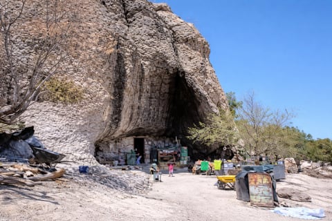 Large rock cave with visitors and vendor stalls in sandy beach setting under clear blue sky