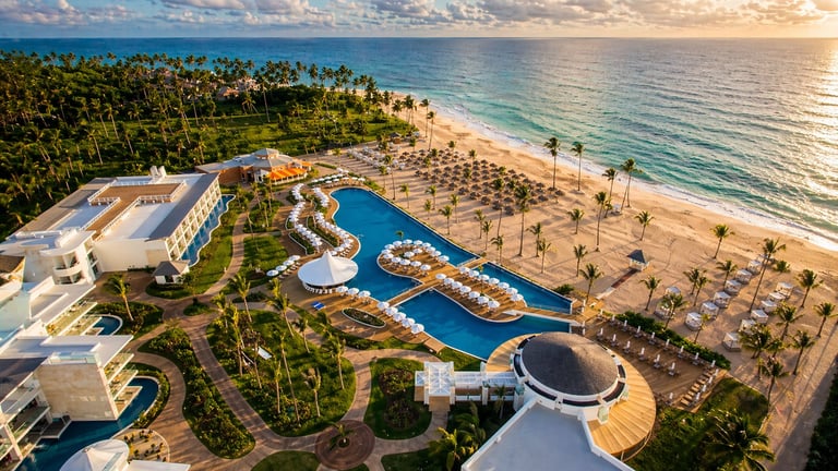Vista aérea del resort frente a la playa con edificios curvos, piscinas azules, palmeras y atardecer sobre el océano