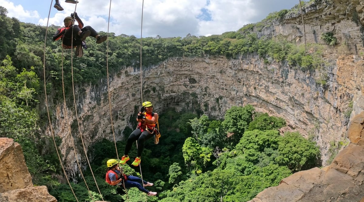 Rock climbers rappelling down a deep sinkhole surrounded by lush green forest and steep limestone cliffs