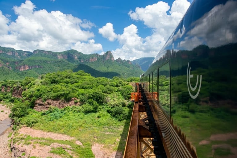 Train traveling through lush green mountains and valleys under a blue sky with white clouds