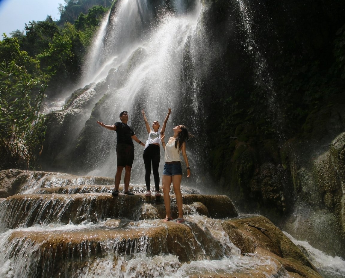 Three people standing on rocks with arms raised in front of a large cascading waterfall surrounded by lush green vegetation