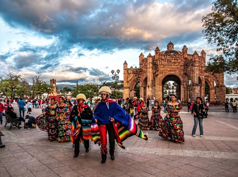 Colorful performers in traditional Mexican attire dance in front of an ornate colonial stone monument under a dramatic cloudy sky