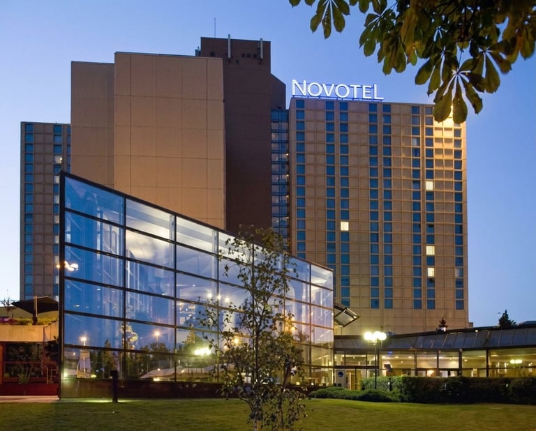 Modern Novotel hotel building at dusk with illuminated facade, blue-tinted glass entrance, and manicured lawn in foreground
