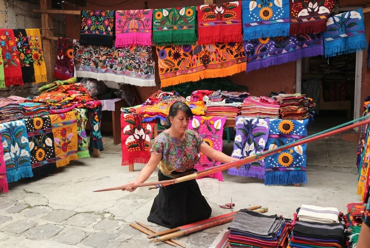 Woman kneeling while weaving on a traditional loom surrounded by colorful textiles and fabrics in an artisan workshop