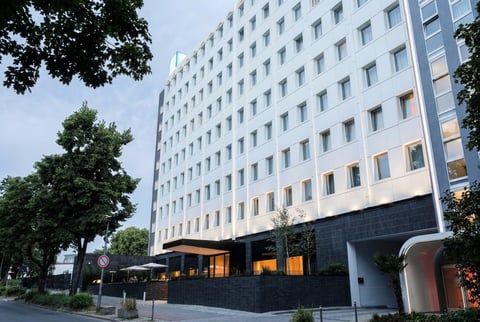 Modern multi-story residential building with white and black facade, illuminated ground floor windows, surrounded by green trees