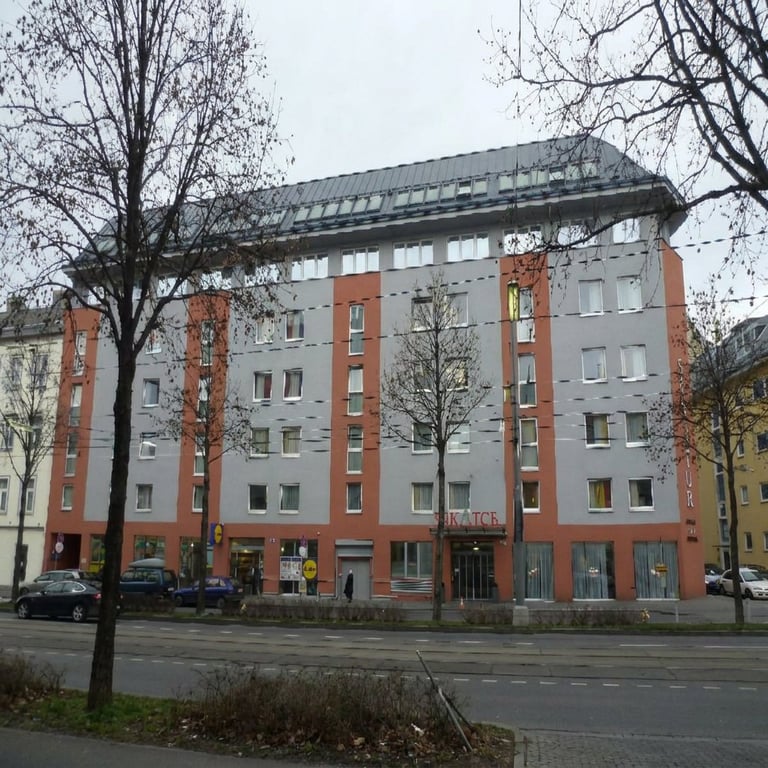 Mid-rise residential apartment building with red brick and gray exterior, bare trees in front, parked cars visible on street