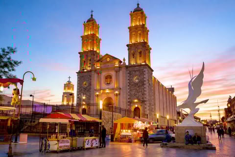 Illuminated church with twin towers at dusk, street vendors and pedestrians in foreground, large bird sculpture to the right.