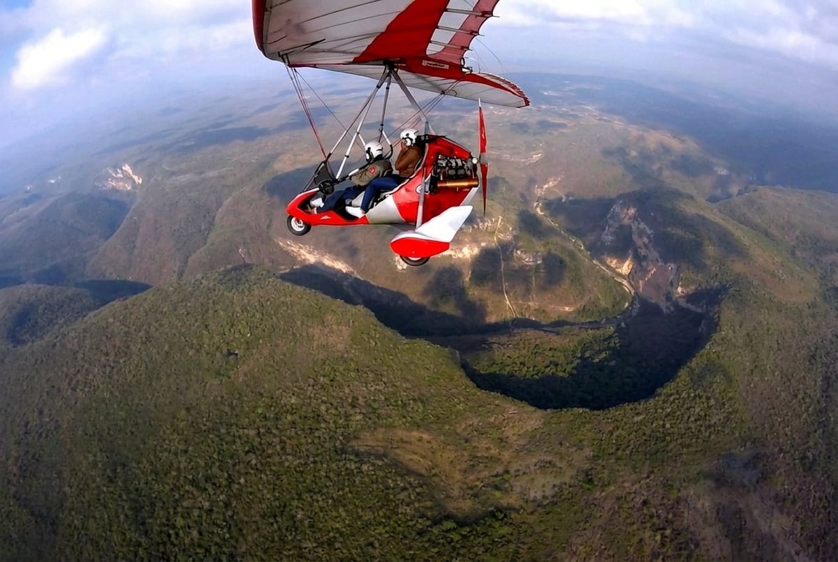 Red and white powered hang glider soaring over forested landscape with lakes and mountains below