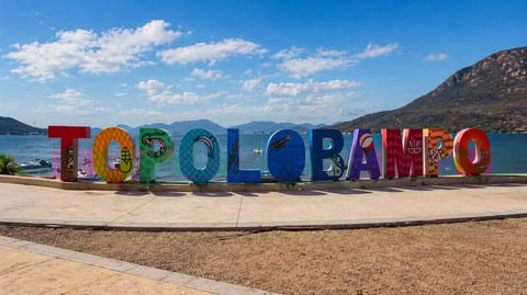 Colorful TOPOLOBAMPO sign on beach with blue water, mountains, and sky in background