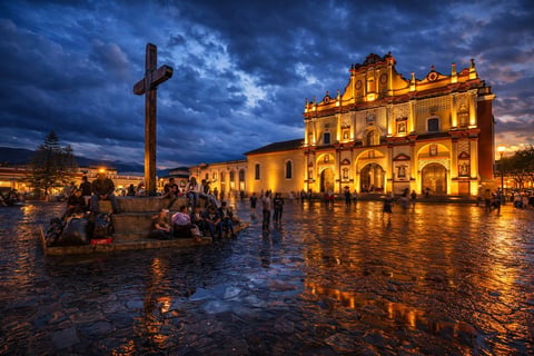 Iglesia colonial iluminada al atardecer con cruz grande en plaza del pueblo, gente reunida, cielo azul tormentoso arriba