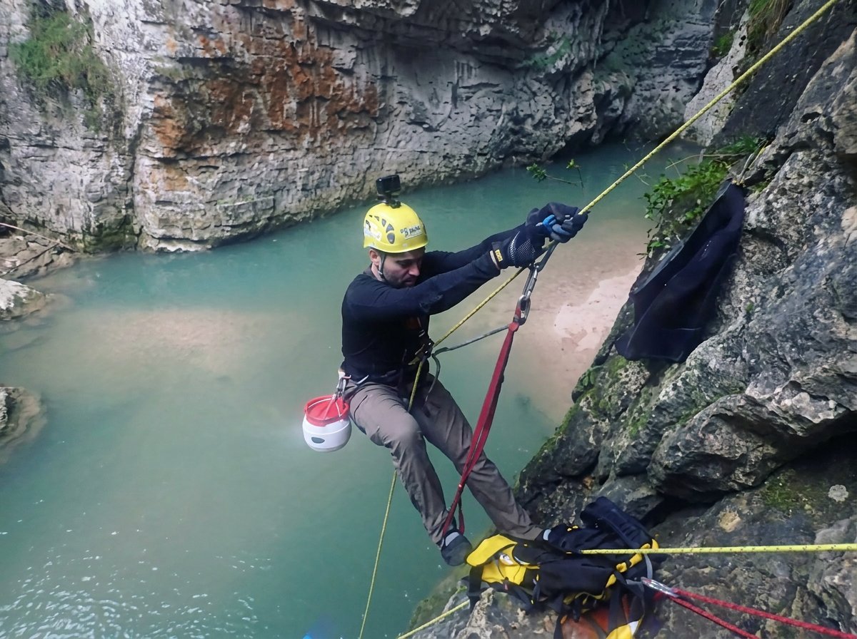 Climber rappelling down a rocky cliff above turquoise water, wearing yellow helmet and safety gear
