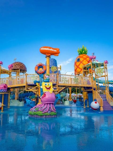 Nickelodeon themed water park playground with colorful structures including a giant pineapple and octopus statue on a blue splash pad