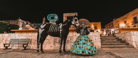 Woman in colorful turquoise dress posing with saddled horse on stone plaza at night with illuminated colonial buildings
