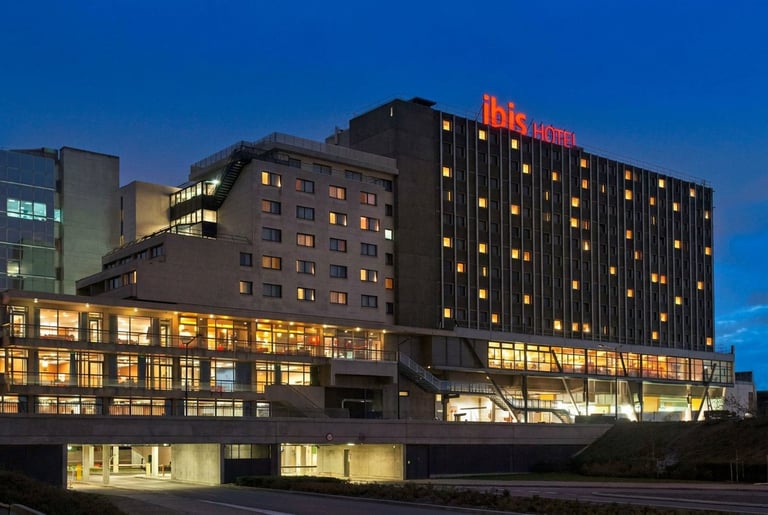 Edificio del hotel Ibis iluminado al anochecer con arquitectura moderna, ventanas iluminadas y señalización roja contra el cielo azul nocturno