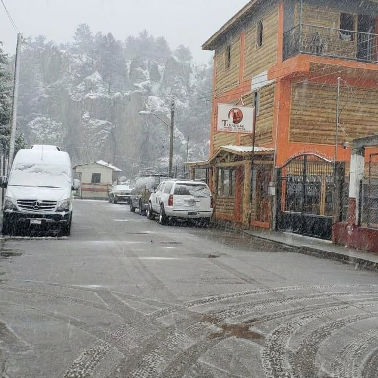 Snowy mountain village street with parked vehicles, rustic buildings, and snow-covered trees in background
