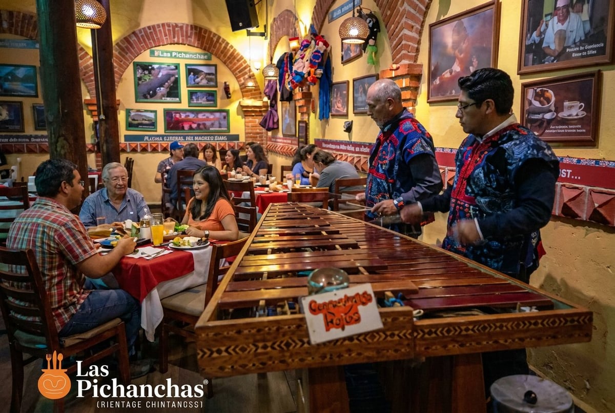 Musicians performing traditional Mexican music on a large wooden guitar-like instrument in a colorful cantina decorated with artwork and memorabilia
