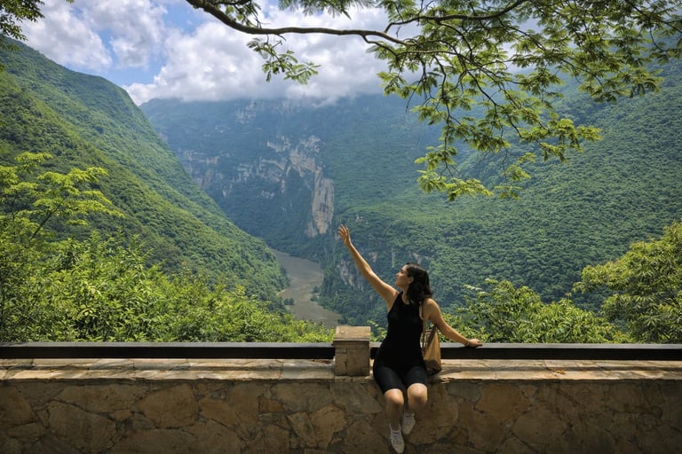 Mujer sentada en un muro de piedra contemplando un exuberante valle verde con montañas y una cascada en la distancia