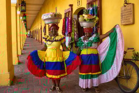 Two women in colorful traditional dresses and headscarves stand in a yellow-columned arcade passageway, displaying draped fabric in the colors of the Colombian flag.