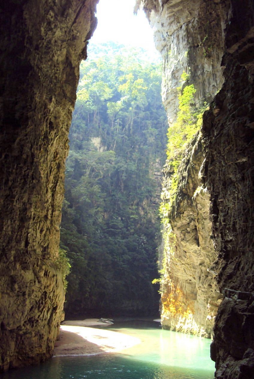 Turquoise river flowing through tall limestone cliffs with lush green forest visible above the canyon opening