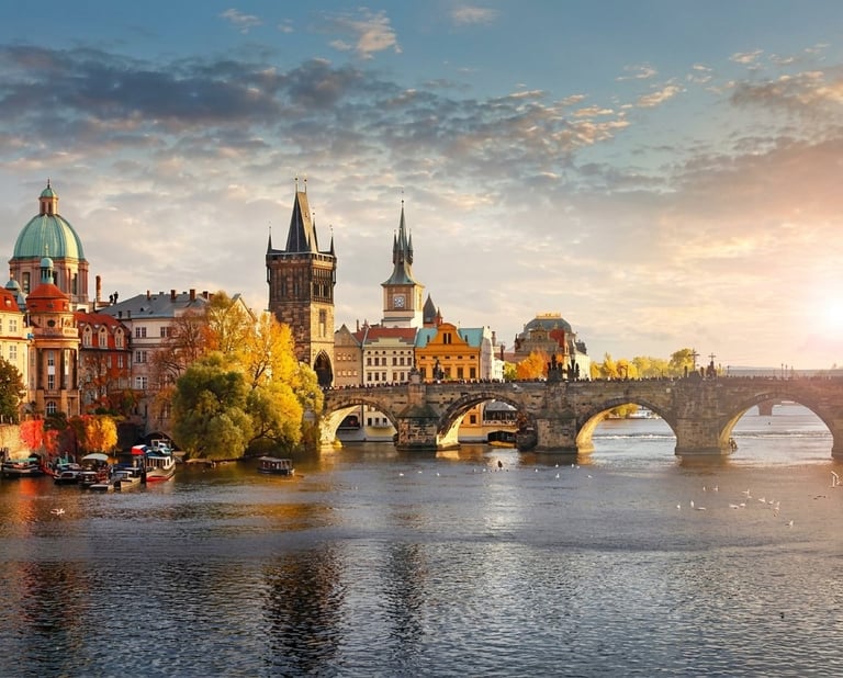 Charles Bridge spanning the Vltava River in Prague at sunset, with historic architecture and spires visible along the riverbank