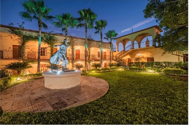 Illuminated Mediterranean-style courtyard at dusk with central sculpture, palm trees, and arched architecture