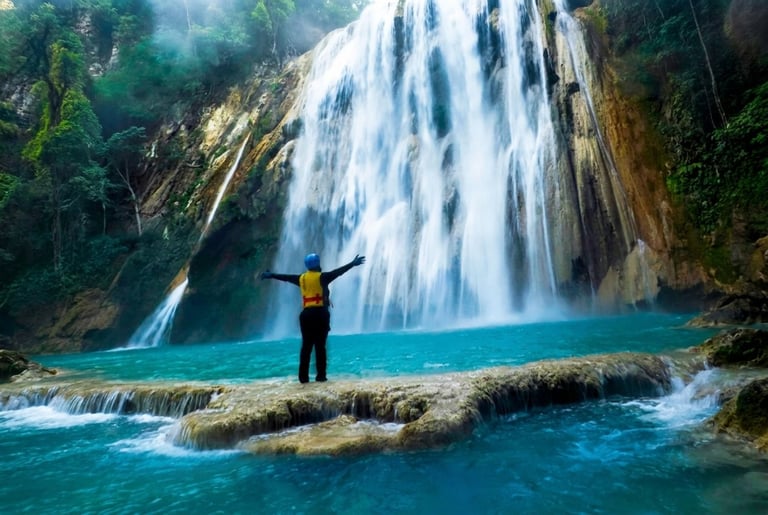 Person with arms raised standing before a large waterfall cascading into turquoise water surrounded by lush green forest