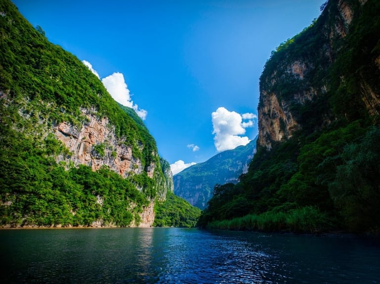 Scenic gorge with steep green mountain cliffs on either side, blue water below, and mountain peak visible in distance under clear sky.