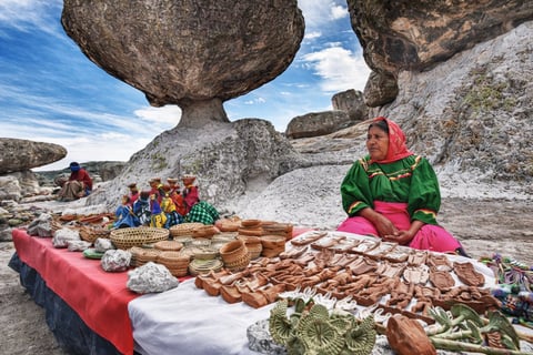 Woman in green and pink clothing displays pottery and crafts on table beneath large balanced boulder rock formation with tourists visiting