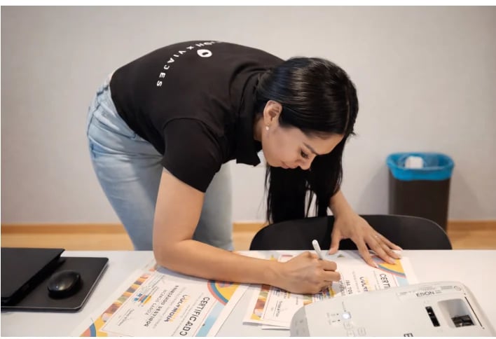 Woman in black shirt reviewing documents at desk with office supplies and blue container in background