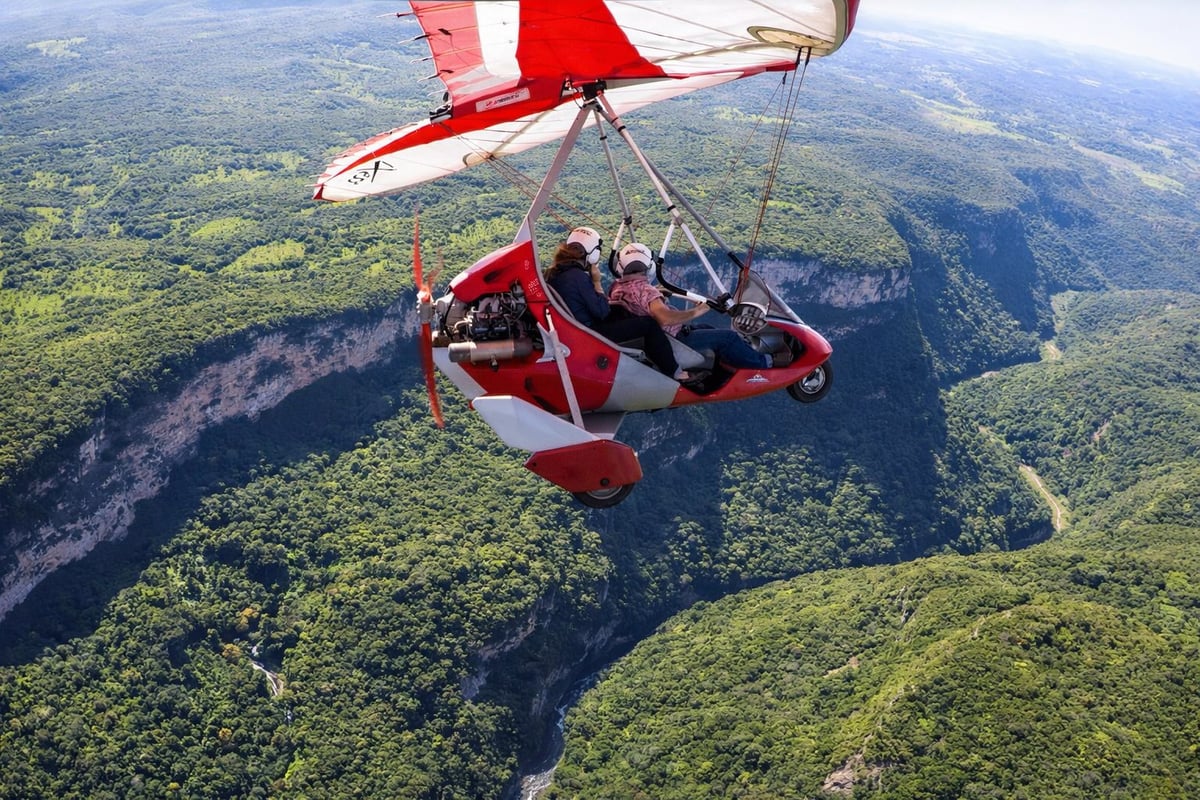 People hang gliding in a red and white aircraft over a forested landscape with a river gorge below
