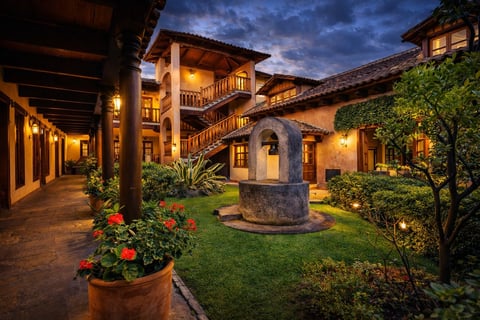 Patio colonial español al atardecer con edificios ocre iluminados, cesped bien cuidado, fuente de piedra y flores en macetas