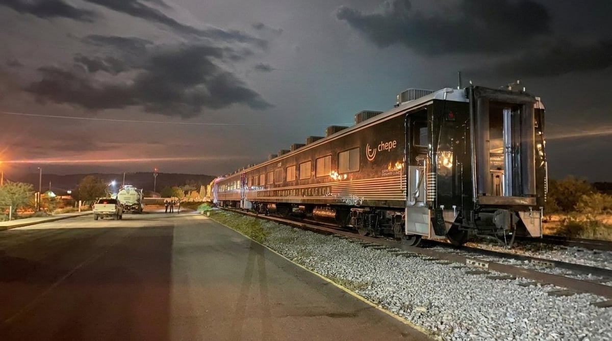 Illuminated passenger train at dusk with golden lighting against a dramatic cloudy sky and gravel track